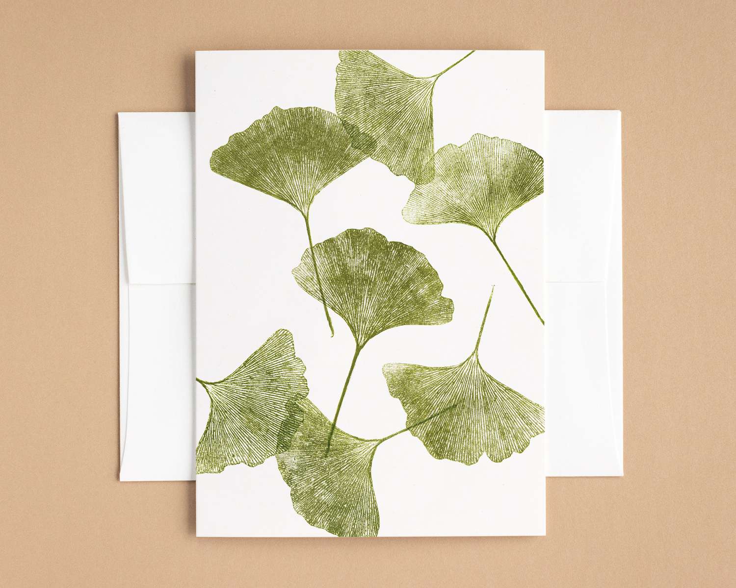 Seven green ginkgo leaves are scattered across the surface of a vertical white greeting card. The card sits on top of a white envelope, which lies on a brown backdrop.